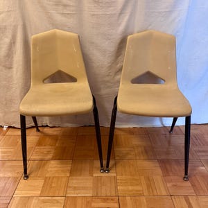 May include: Two beige plastic chairs with black metal legs. Each chair has a triangular cutout near the top of the backrest. The chairs are positioned on a wooden floor.