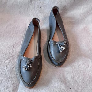 May include: A pair of gray leather loafers with decorative tassels. The shoes have a rounded toe and a stitched design around the edges. The loafers are displayed on a neutral-colored fabric background.