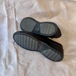 Op de afbeelding: Een paar zwarte Clarks-schoenen, gezien vanaf de zolen. De zolen zijn donkergrijs met een geribbeld patroon voor grip. De merknaam "Clarks" is zichtbaar op de zool. De schoenen liggen op een lichtgekleurd, getextureerd oppervlak.