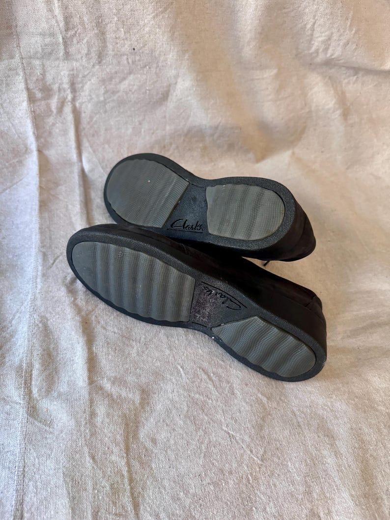 Op de afbeelding: Een paar zwarte Clarks-schoenen, gezien vanaf de zolen. De zolen zijn donkergrijs met een geribbeld patroon voor grip. De merknaam "Clarks" is zichtbaar op de zool. De schoenen liggen op een lichtgekleurd, getextureerd oppervlak.