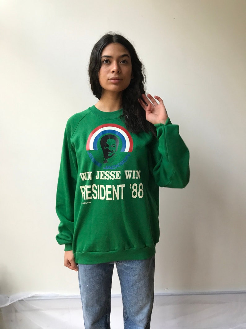 Puede incluir: Una sudadera verde de manga larga con un gr&aacute;fico de Jesse Jackson bajo un arco&iacute;ris rojo, blanco y azul. El texto dice RUN JESSE WIN PRESIDENT '88 en letras blancas. La sudadera tiene cuello redondo y pu&ntilde;os acanalados.