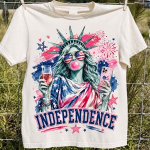Könnte beinhalten: Cremefarbenes T-Shirt mit einer Grafik der Freiheitsstatue mit Sonnenbrille und Kaugummiblase, die ein Getränk und ein Telefon hält. Das Wort "INDEPENDENCE" ist unten in Rot und Blau aufgedruckt.