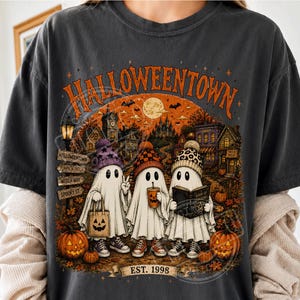 May include: A charcoal gray t-shirt featuring a graphic print of three ghosts wearing beanies, holding a pumpkin bag, a coffee cup, and a book. The text reads HALLOWEENTOWN and EST. 1998, with a background of a spooky town, pumpkins, and a full moon.