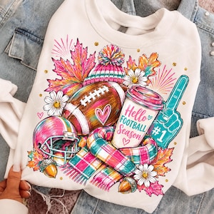 May include: A white crewneck sweatshirt featuring a colourful graphic print of a football, a pink helmet, a coffee cup with the text Hello FOOTBALL Season, a foam finger with #1, a plaid scarf, autumn leaves, daisies, and acorns.