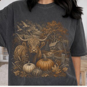 May include: A charcoal gray short-sleeve t-shirt featuring a detailed graphic print of a Highland cow standing in a field with pumpkins, a wooden fence, a barn, trees, and flying birds in a warm, rustic, earth-toned color palette.