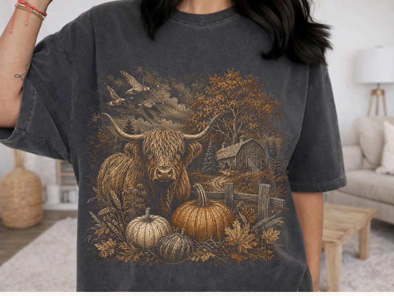 May include: A charcoal gray short-sleeve t-shirt featuring a detailed graphic print of a Highland cow standing in a field with pumpkins, a wooden fence, a barn, trees, and flying birds in a warm, rustic, earth-toned color palette.