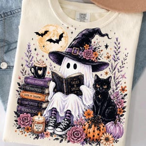 May include: A cream-colored t-shirt featuring a graphic of a ghost wearing a witch hat and reading a book titled Tales & Spells. The design includes a stack of books labeled Magic Potions, Enchantments, Book of Shadows, Love Spells, and Herbology.