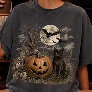 May include: A charcoal gray t-shirt featuring a detailed graphic print of a jack-o'-lantern pumpkin, a black cat sitting in tall grass, a bare tree, and bats flying in front of a large full moon against a dark, atmospheric night sky.