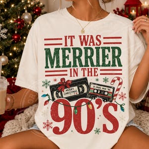 May include: An off-white t-shirt featuring the text "IT WAS MERRIER IN THE 90'S" in red and green. The graphic includes a black VHS tape with a "MERRY CHRISTMAS" label, a cassette tape labeled "X-MAS MIX '95", a candy cane, and string lights.
