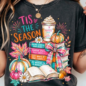 May include: A charcoal gray t-shirt featuring a colorful graphic of a stack of books, a pumpkin spice latte, pumpkins, and autumn leaves. The text reads 'Tis The Season', 'Pumpkin Spice & Good Books', and various book-themed phrases.