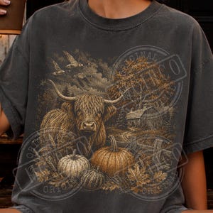 Puede incluir: Una camiseta gris carbón con un estampado gráfico detallado de una vaca de las Tierras Altas en un campo con calabazas, una valla de madera, un granero y pájaros volando en el cielo. El diseño utiliza tonos marrones y dorados terrosos.
