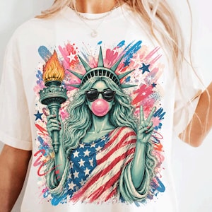 Könnte beinhalten: Cremefarbenes T-Shirt mit einer Grafik der Freiheitsstatue, die eine Sonnenbrille trägt und eine Kaugummiblase bläst. Die Freiheitsstatue ist in eine amerikanische Flagge gehüllt und zeigt das Friedenszeichen. Bunte Farbspritzer und Sterne vervollständigen das Design.