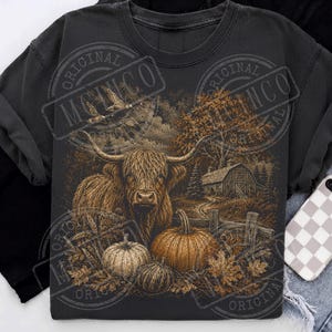May include: A charcoal gray crewneck t-shirt featuring a graphic print of a Highland cow standing in a field with pumpkins, a wooden fence, and a barn in the background. The design includes autumn leaves and flying birds in a sepia-toned illustration.
