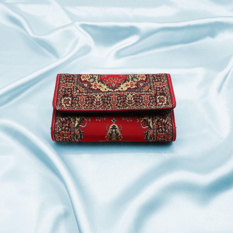 May include: A red wallet with a patterned design resembling a rug. The pattern is in shades of black, gold, and red.