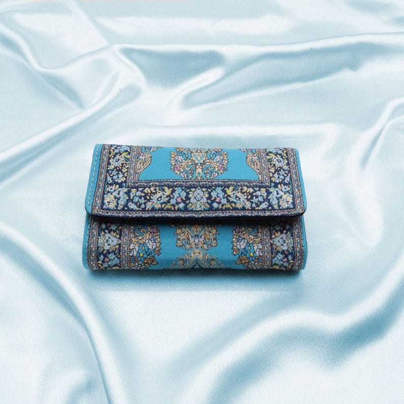 May include: A blue wallet with a patterned design that resembles a rug. The wallet is folded closed and sitting on a light blue satin surface.