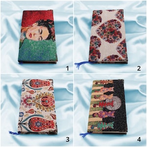 May include: Four different fabric-covered notebooks with colorful patterns. The notebooks are all closed and have a blue ribbon bookmark. The patterns include a portrait of Frida Kahlo, a floral design, a geometric design, and a design with cats.