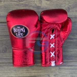Puede incluir: Un par de guantes de boxeo rojos con cordones blancos. Los guantes tienen el texto "NO BOXING NO LIFE" en la muñeca. Están hechos de un material brillante y se muestran sobre una superficie de madera.