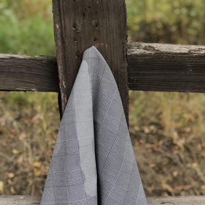 Pu&ograve; includere: Un tessuto a quadri grigio e bianco drappeggiato su un palo di recinzione in legno stagionato. Il tessuto presenta un classico motivo a quadri nelle tonalit&agrave; del grigio e del bianco. Lo sfondo &egrave; sfocato, suggerendo un ambiente esterno.