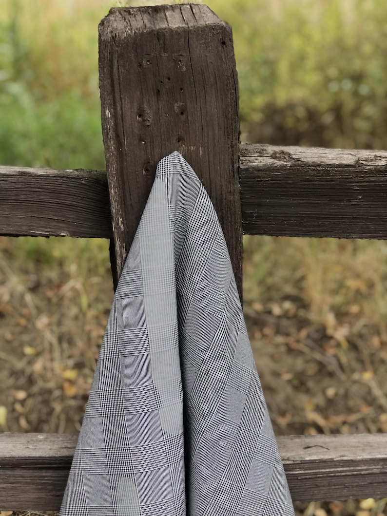 Pu&ograve; includere: Un tessuto a quadri grigio e bianco drappeggiato su un palo di recinzione in legno stagionato. Il tessuto presenta un classico motivo a quadri nelle tonalit&agrave; del grigio e del bianco. Lo sfondo &egrave; sfocato, suggerendo un ambiente esterno.