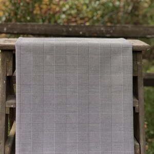 Pu&ograve; includere: Un tessuto a quadri grigio e bianco drappeggiato su una superficie di legno. Il tessuto presenta un classico motivo a quadri, con sottili linee bianche che si intersecano su uno sfondo grigio. La superficie in legno &egrave; un piccolo tavolo o una panca, con finitura in legno naturale.