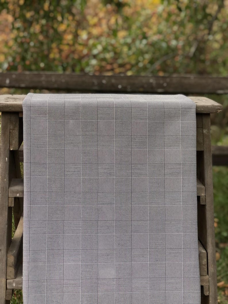 Pu&ograve; includere: Un tessuto a quadri grigio e bianco drappeggiato su una superficie di legno. Il tessuto presenta un classico motivo a quadri, con sottili linee bianche che si intersecano su uno sfondo grigio. La superficie in legno &egrave; un piccolo tavolo o una panca, con finitura in legno naturale.