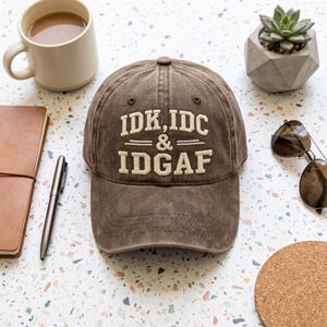May include: A brown baseball cap with a distressed finish featuring the embroidered cream-colored text IDK, IDC & IDGAF on the front panel. The cap has a structured crown, a curved brim, and stitched eyelets for ventilation on the top panels.
