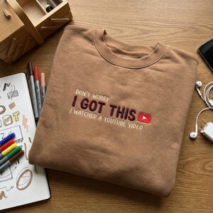 May include: A brown sweatshirt with the embroidered text "DON'T WORRY I GOT THIS I WATCHED A YOUTUBE VIDEO" in white and red. The YouTube logo is also embroidered in red. The sweatshirt is folded on a wooden surface.