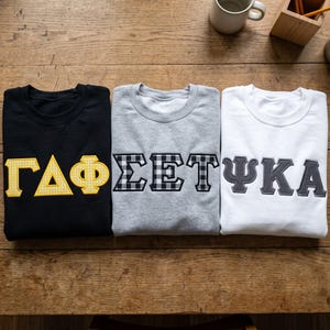 Könnte beinhalten: Drei Rundhals-Sweatshirts in Schwarz, meliertem Grau und Weiß. Das schwarze Sweatshirt hat gelbe, karierte griechische Buchstaben. Das graue Sweatshirt zeigt griechische Buchstaben mit schwarz-weißem Karomuster. Das weiße Sweatshirt hat dunkelgraue griechische Buchstaben.