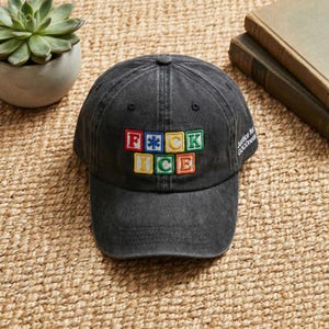 May include: A dark grey baseball cap featuring the embroidered phrase "F*CK ICE" in colourful block letters. The cap also has the text "Justice for GOODness" on the side. The hat is placed on a woven surface.