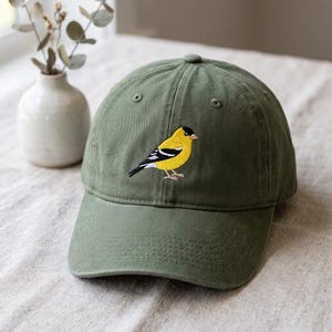 Könnte beinhalten: Eine olivgrüne Baseballkappe mit einer Stickerei eines Goldzeisigs auf der Vorderseite. Der Vogel ist mit leuchtend gelben Federn, schwarzen Flügeln mit weißen Flügelbinden und einer schwarzen Kappe auf dem Kopf über dem Schirm dargestellt.