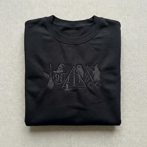 May include: Black crew neck sweatshirt featuring embroidered symbols: a broom, sorting hat, owl, Deathly Hallows symbol, wizard, and glasses. The symbols are tone-on-tone.