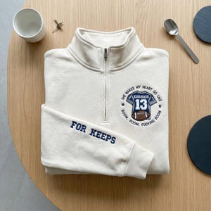 May include: Cream-colored quarter-zip sweatshirt with navy blue embroidered text and graphics. The left sleeve reads "FOR KEEPS." The chest features a football jersey design with the name "KAVANAGH" and the number "13." The design includes the phrase "SHE MAKES MY HEART GO LIKE BOOM, BOOM, FUCKING BOOM."