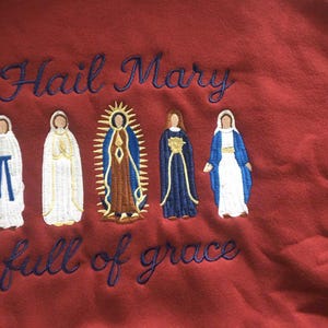 May include: Maroon sweatshirt with embroidered figures of religious figures. The text "Hail Mary full of grace" is embroidered in blue script above and below the figures. The figures are in white, brown, and blue.