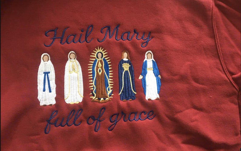 May include: Maroon sweatshirt with embroidered figures of religious figures. The text "Hail Mary full of grace" is embroidered in blue script above and below the figures. The figures are in white, brown, and blue.