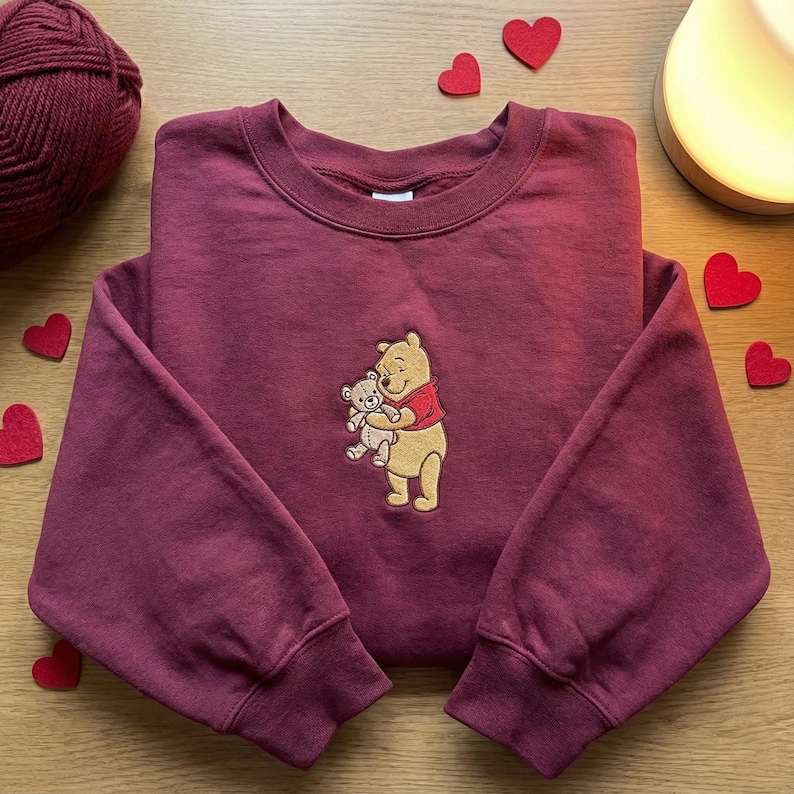 May include: A maroon sweatshirt featuring an embroidered image of Winnie the Pooh hugging a teddy bear. The sweatshirt has long sleeves and a crew neck. Red heart-shaped decorations are scattered around the sweatshirt.