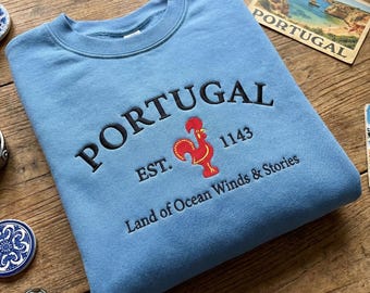 May include: A light blue sweatshirt with the word "PORTUGAL" embroidered in black, along with a red rooster emblem and the text "EST. 1143". The phrase "Land of Ocean Winds & Stories" is also embroidered on the sweatshirt.