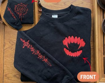 May include: Black sweatshirt with red embroidered designs. The front features a stylized fangs design, while the back has a circular pattern with vertical lines. The left sleeve displays a sword design.