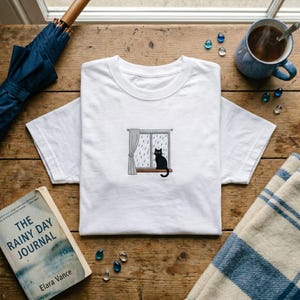 May include: White t-shirt with an embroidered design of a black cat looking out of a window in the rain. The shirt is on a wooden surface, alongside a book titled "The Rainy Day Journal" and a blue umbrella.