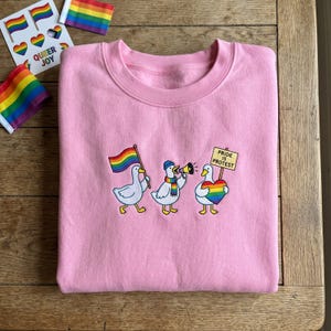 May include: A light pink crewneck sweatshirt featuring an embroidered graphic of three white ducks. One duck carries a rainbow flag, another wears a rainbow scarf and holds a megaphone, and the third holds a rainbow heart and a sign reading PRIDE IS PROTEST.