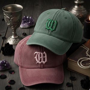 May include: Two baseball caps, one dusty rose and one green, are displayed. Each cap features an embroidered letter "W" in a contrasting color. The caps are on a wooden surface with gothic-themed accessories, including a chalice and crystals.