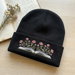 May include: Black beanie hat with an embroidered design of an open book with pink flowers and green leaves. The hat is made of a soft, knitted material and is suitable for cold weather.