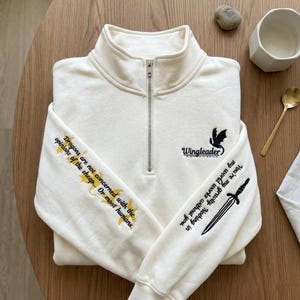 May include: Off-white quarter-zip sweatshirt with embroidered text and graphics. The left sleeve has text and gold star accents. The right sleeve features a sword graphic and text. The chest displays a dragon graphic and the word "Wingleader".