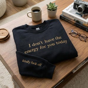 Pode incluir: Camisola preta com texto dourado que diz "I don't have the energy for you today" e "kindly fuck off". A camisola está numa mesa de madeira com uma chávena de café, uma planta e livros. Também são visíveis óculos e uma câmara.