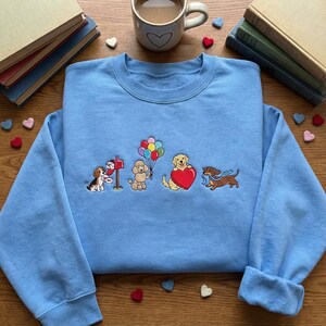 Peut inclure: Un sweat-shirt bleu clair avec des chiens brodés. Un beagle livre le courrier, un caniche tient des ballons, un golden retriever tient un cœur et un teckel porte une écharpe. Livres et une tasse à café avec des détails de cœur en arrière-plan.