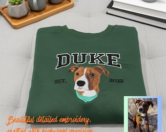 May include: A dark green sweatshirt with the name "DUKE" embroidered in white and black lettering. The sweatshirt features an embroidered dog portrait with a teal scarf, the text "EST. 2022", and a small photo of the dog.