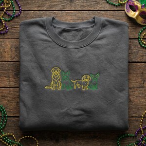 Peut inclure: Un sweat-shirt gris foncé avec une broderie de quatre chiens en jaune et vert. Les chiens sont disposés en rang, avec un golden retriever, un bouledogue français, un teckel et un chihuahua. Le sweat-shirt est plié sur une surface en bois.