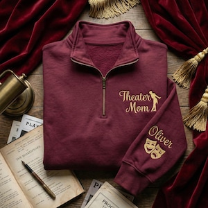 May include: A burgundy quarter-zip sweatshirt with gold embroidered text and graphics. The text reads "Theater Mom" with a silhouette of a person. The sleeve has the name "Oliver" and theater masks.