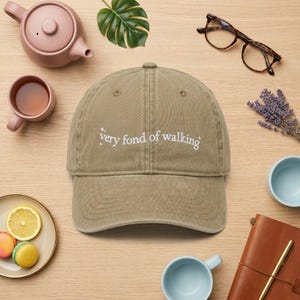 May include: A khaki baseball cap with the embroidered text "very fond of walking". The image includes a teapot, teacup, spectacles, lavender, and macarons, all arranged on a wooden surface.