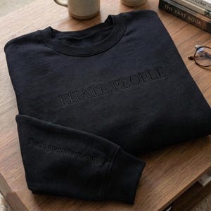 May include: Black sweatshirt with the embroidered text "I HATE PEOPLE" across the chest. The sleeve features the embroidered text "Fuck, people are stupid". The sweatshirt is on a wooden surface.