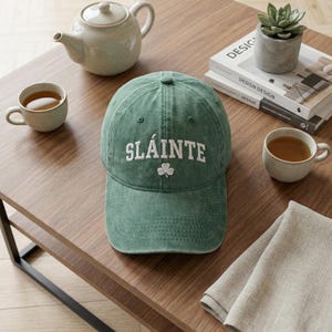 Könnte beinhalten: Eine grüne Baseballkappe mit dem weißen Wort "SLÁINTE" und einem Kleeblatt. Auf einem Holztisch, neben einer Teekanne, Teetassen und einer Sukkulentenpflanze.